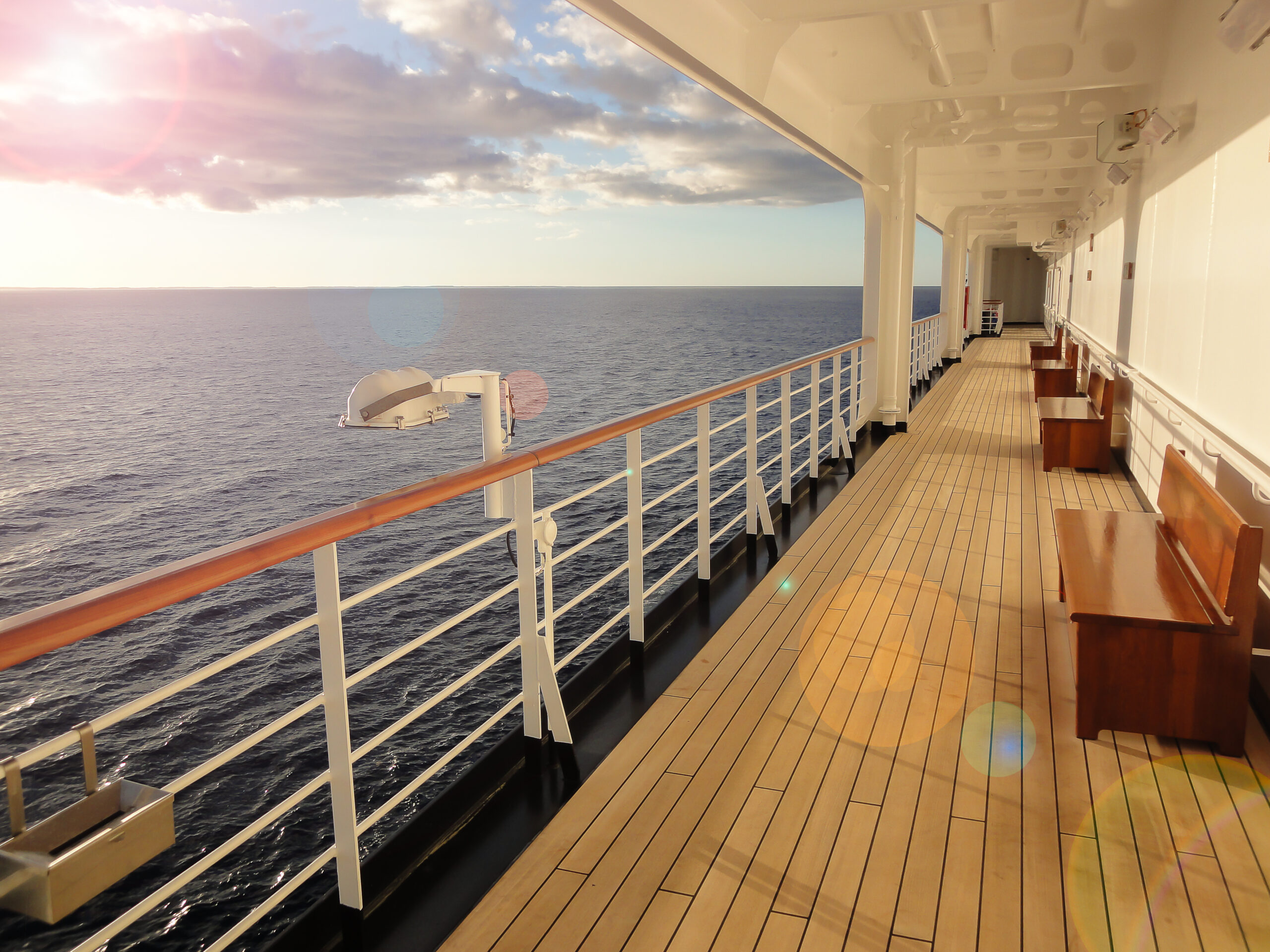 A cruise ship deck with wooden benches and railings overlooking a calm ocean under a partly cloudy sky.