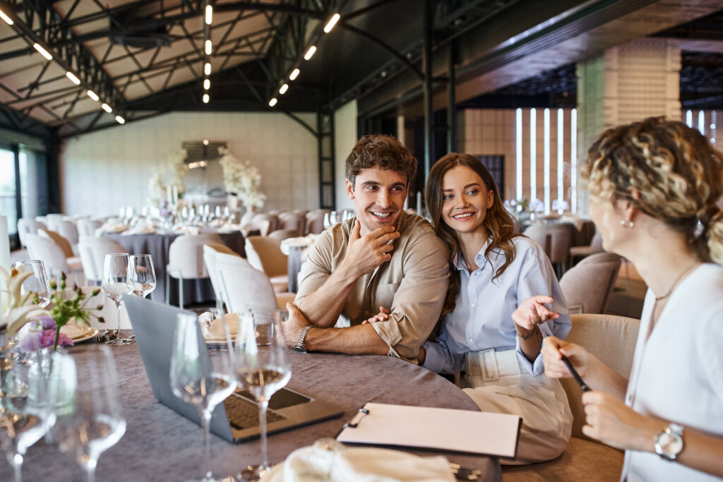 Happy couple and event manager discussing wedding preparation at festive table in banquet hall